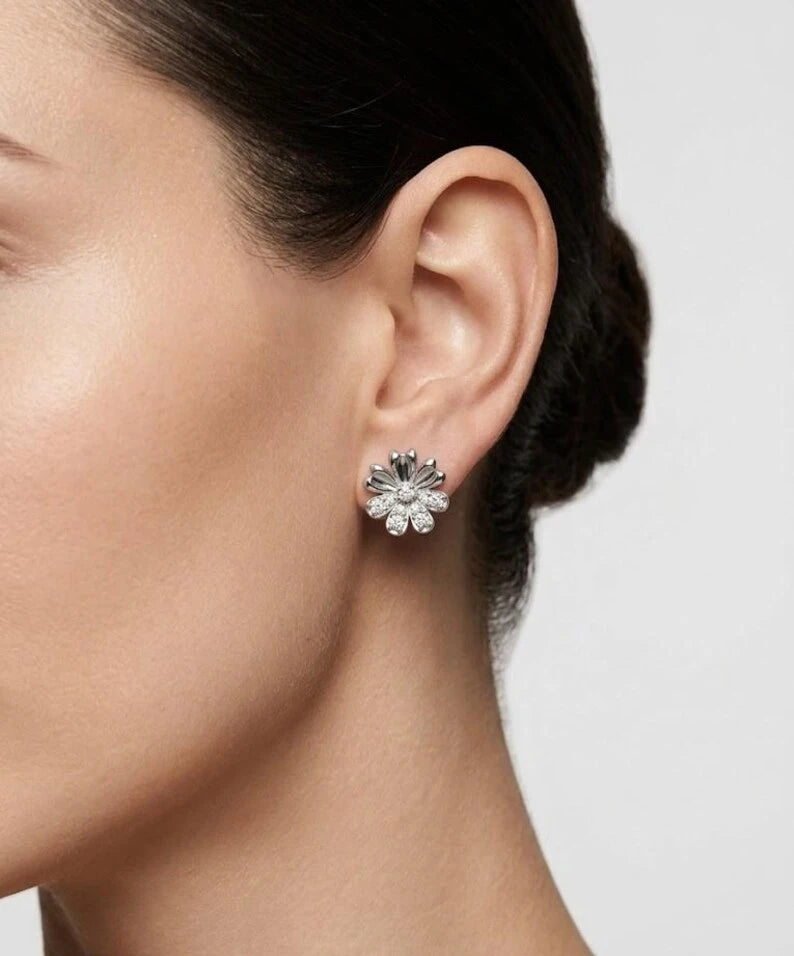 A close-up side profile of a woman wearing a silver-toned flower-shaped stud earring with clear stones, set against a plain light gray background.
