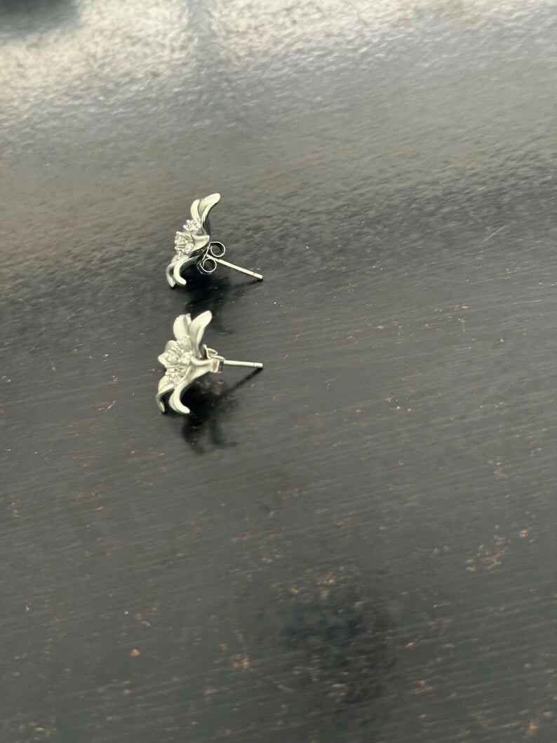 A pair of silver-toned lily flower stud earrings resting on a dark, textured wooden surface.