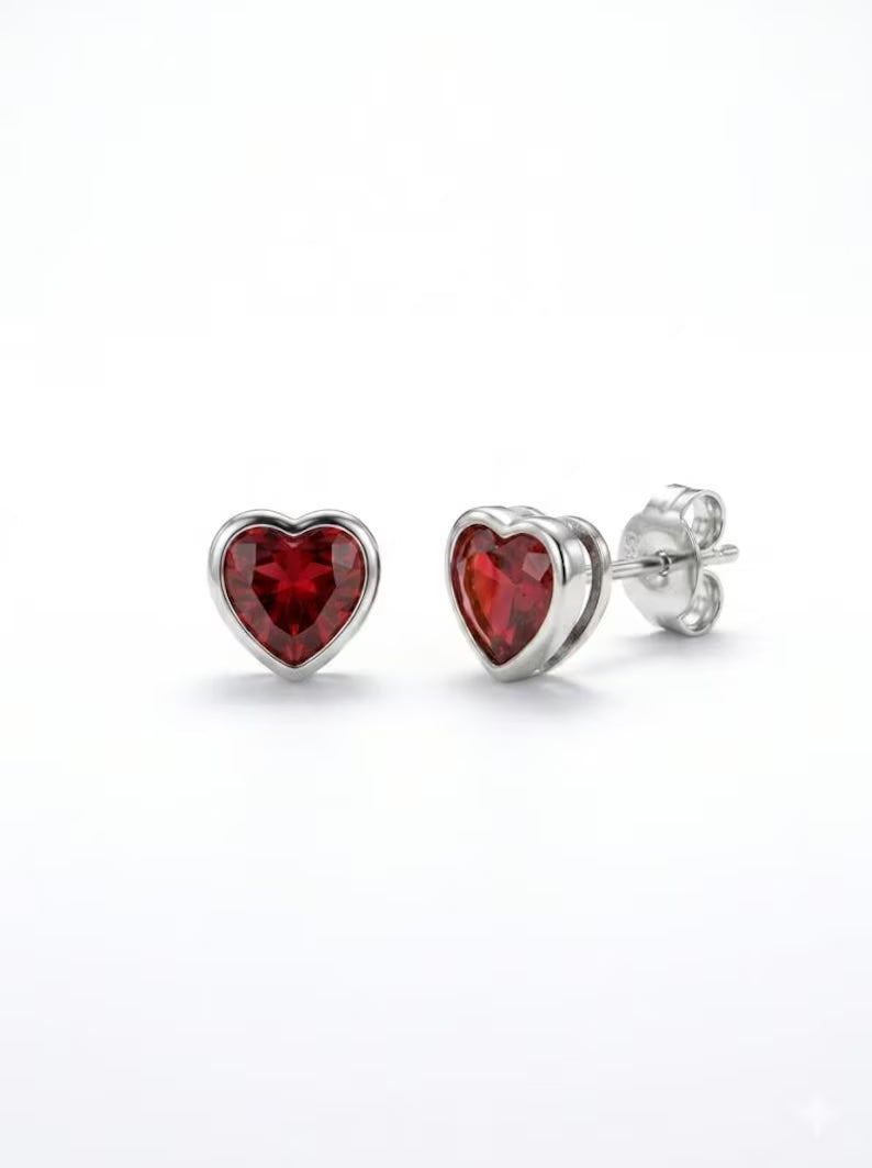 A pair of heart-shaped stud earrings with faceted red gemstones in silver bezel settings, one facing forward and the other angled to show the post and backing, on a clean white background.