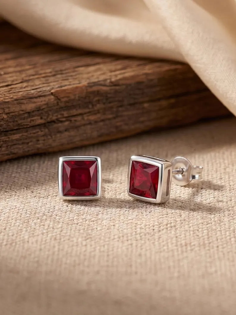 A pair of square stud earrings with deep red gemstones in silver bezel settings, displayed on a beige fabric surface with a rustic wooden prop and draped satin in the background.