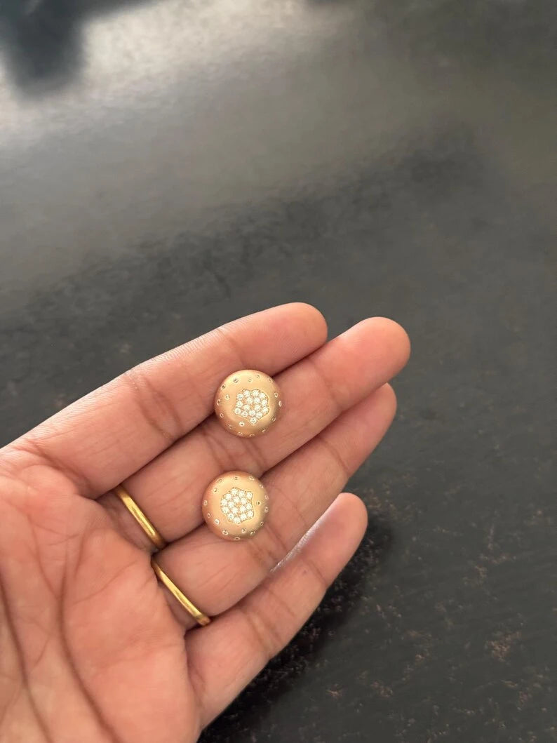 A close-up of a hand holding a pair of small round gold stud earrings with a textured center, set against a dark, out-of-focus background.