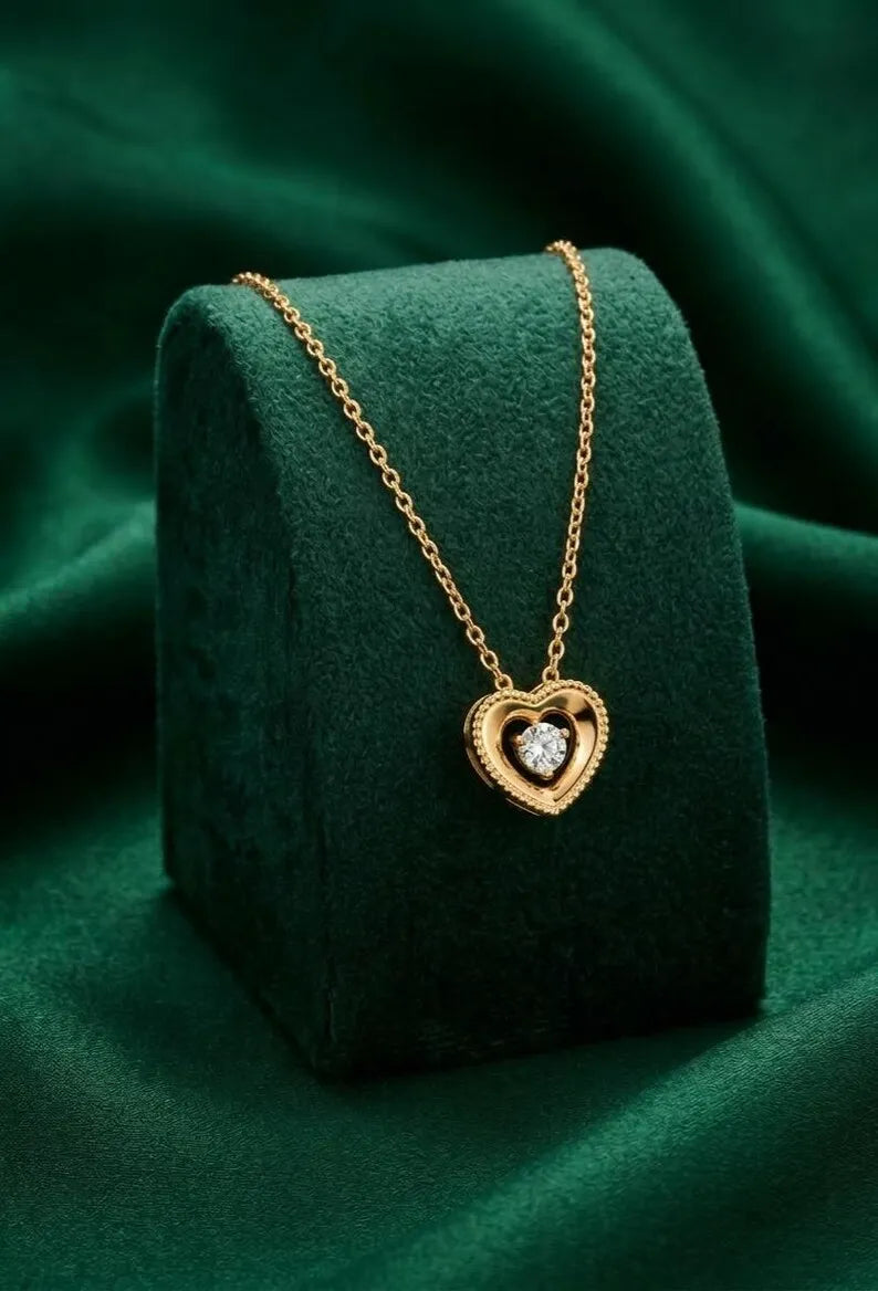 A gold heart pendant necklace with a central clear gemstone, displayed on a dark green velvet jewelry stand against a draped green fabric background.