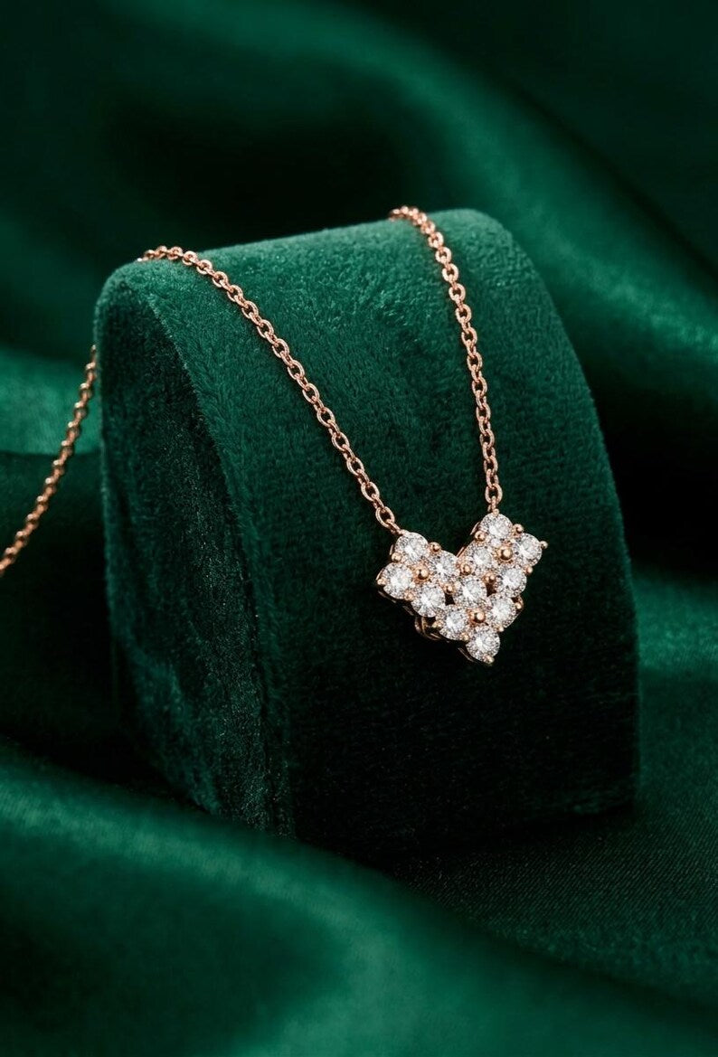 A rose gold necklace with a heart-shaped pendant set with clear gemstones, displayed on a dark green velvet jewelry stand against a matching green fabric background.
