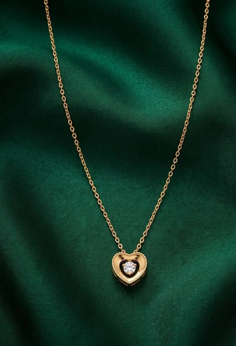 A gold heart pendant necklace with a clear central gemstone, displayed on a dark green satin fabric background.
