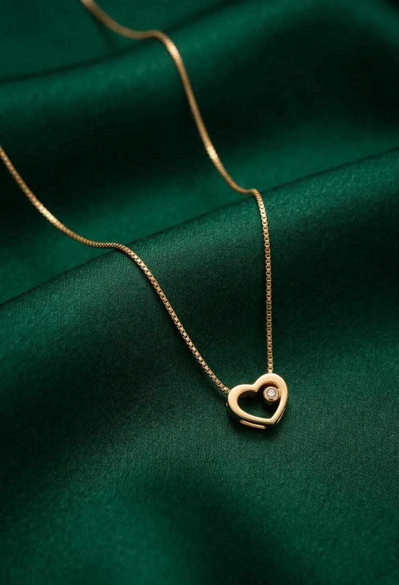 A delicate gold chain necklace with an open heart pendant featuring a small clear gemstone, displayed on a draped dark green satin fabric.