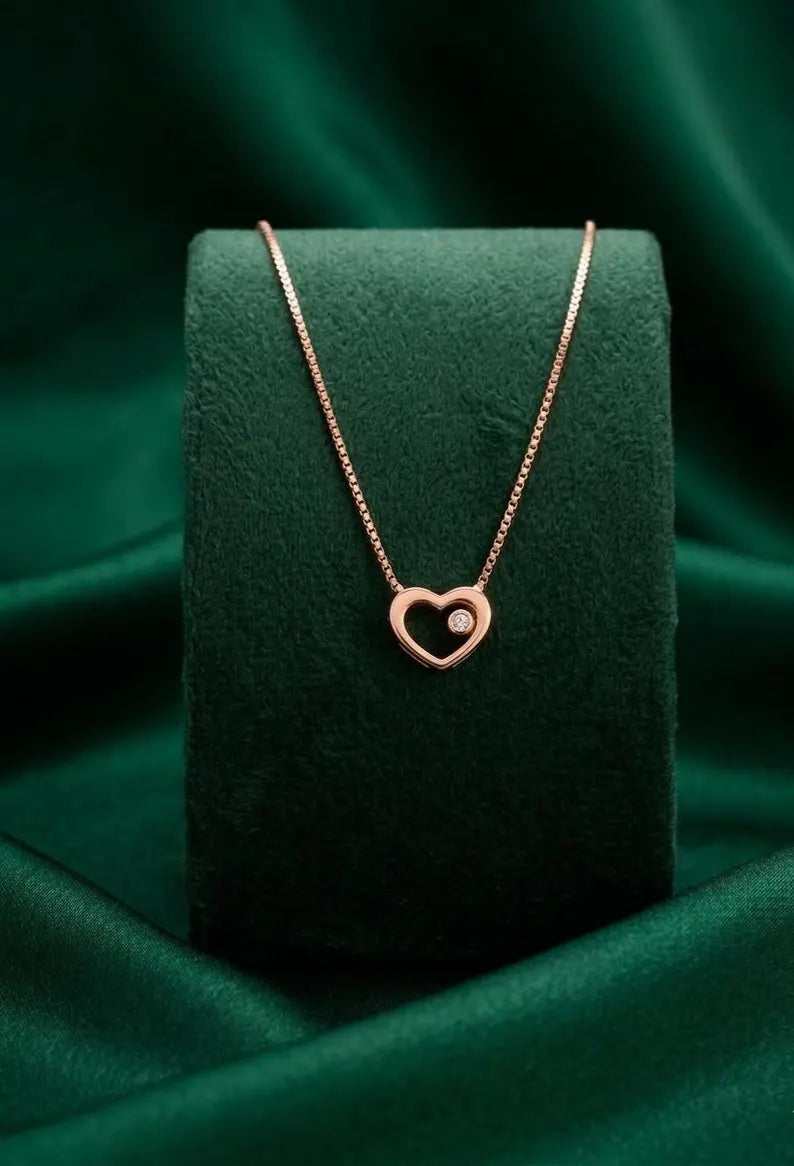 A delicate rose gold necklace with a heart-shaped pendant displayed on a dark green velvet jewelry stand, set against a draped dark green satin fabric background.