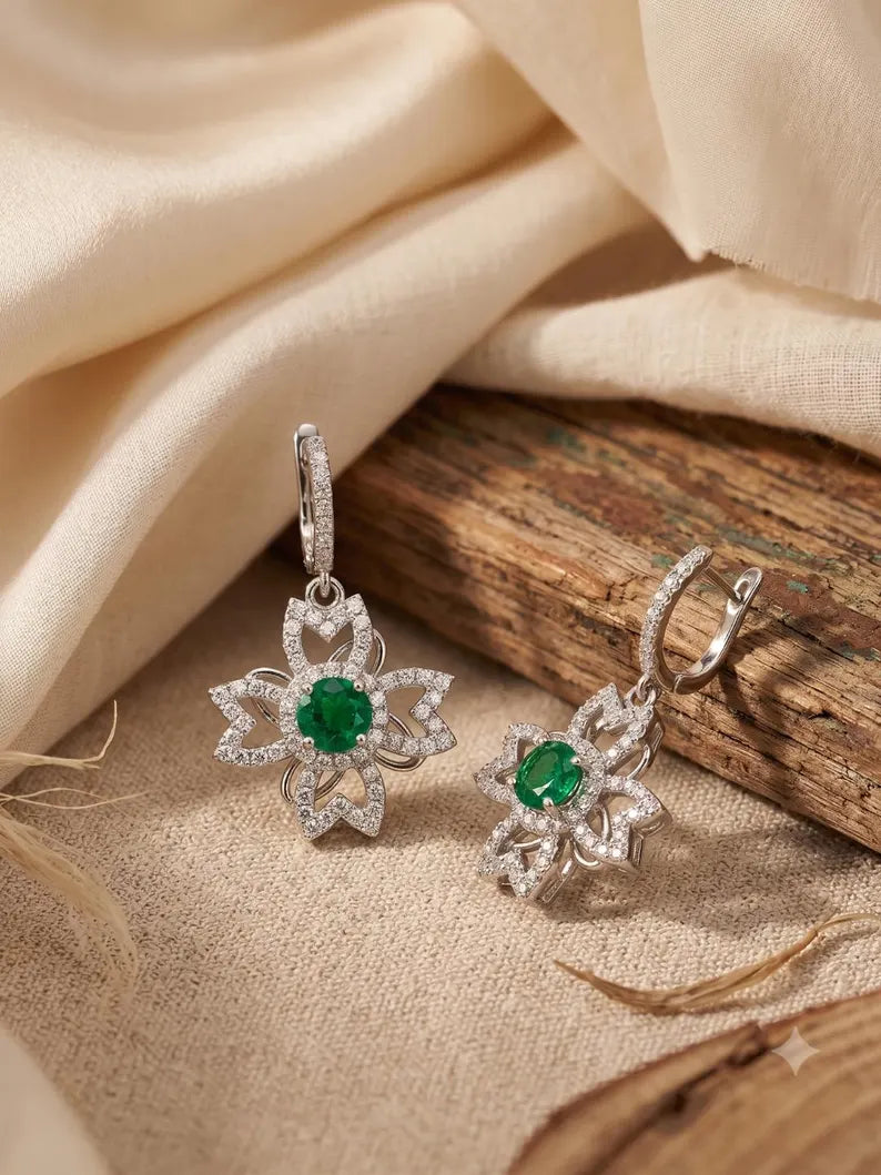 A pair of silver-toned dangle earrings with starburst floral designs, each featuring a round green emerald center stone and pavé clear stones, displayed on a textured beige fabric next to a rustic wooden slice.
