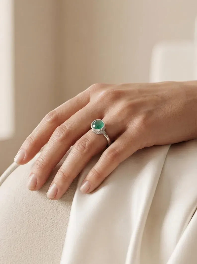 Hand resting on cream satin fabric wearing a minimalist silver ring with a green bezel-set stone