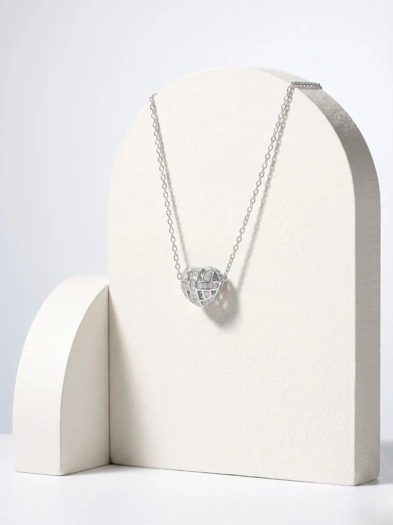 A silver chain necklace with a textured, diamond-like spherical pendant displayed on a cream-colored arched stand against a light gray background.