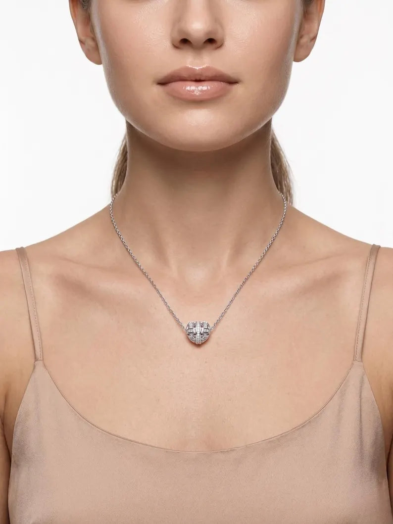 A close-up of a model wearing a delicate silver chain necklace with a small, clear, heart-shaped crystal pendant. The model is wearing a beige camisole top against a clean, light gray background.