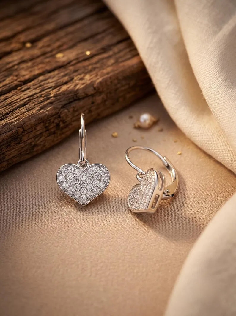 A pair of silver heart-shaped hoop earrings with pavé-set clear stones, displayed on a beige fabric surface next to a rustic wooden block.