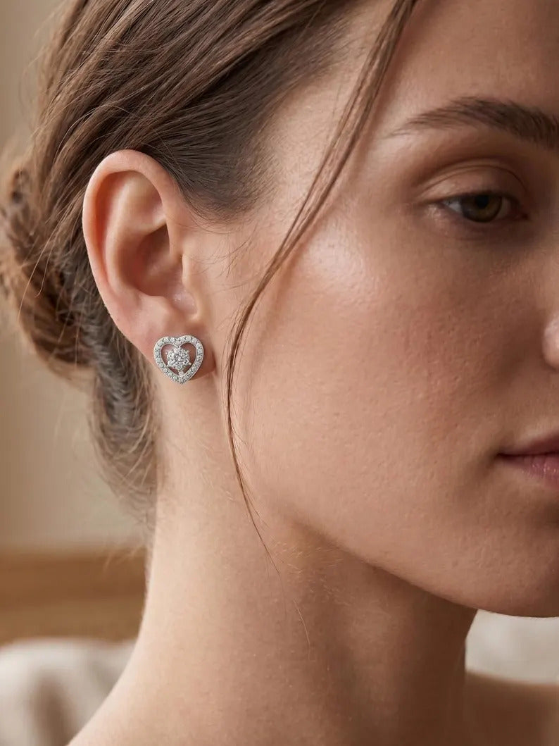 A close-up of a woman's ear wearing a small heart-shaped stud earring with a silver-toned metal border and a central cluster of clear stones.