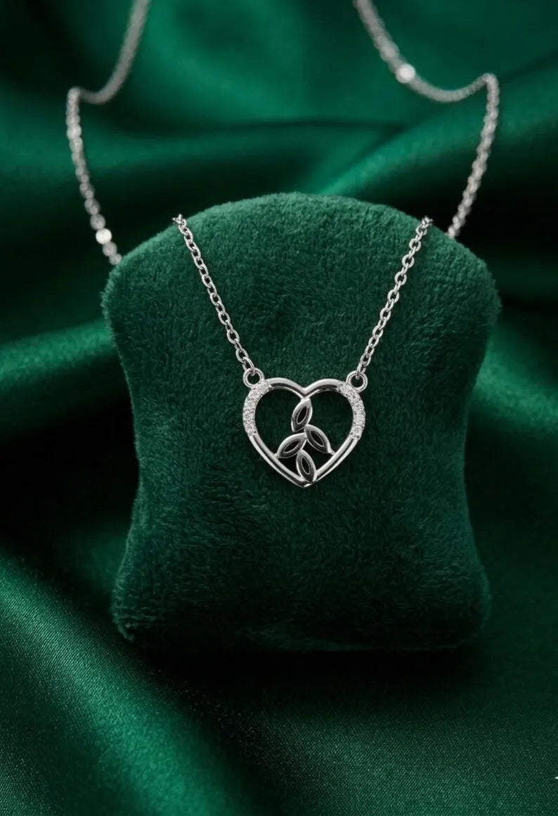 A silver heart-shaped pendant necklace with a Celtic triquetra symbol inside, displayed on a dark green velvet jewelry cushion over a matching green satin fabric background.