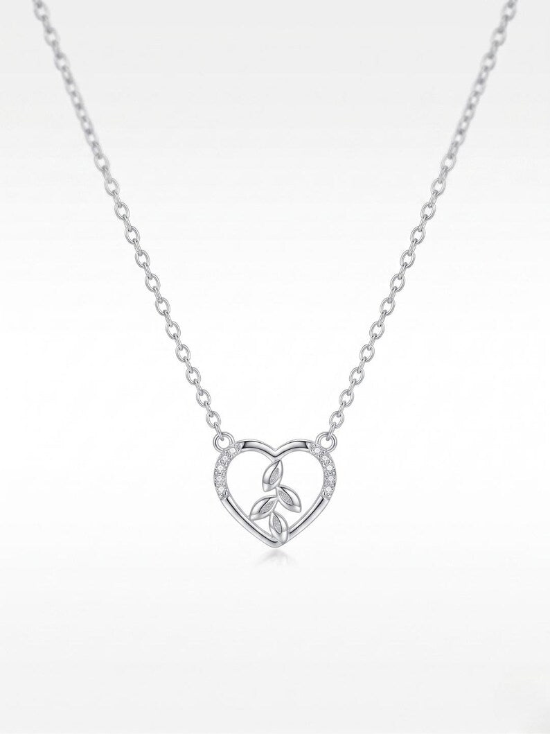 A delicate silver chain necklace featuring a heart-shaped pendant with a central floral motif, photographed against a clean white background.