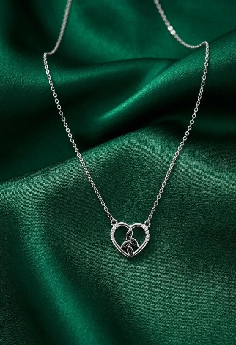 A silver heart-shaped pendant necklace with a small central emblem, displayed on draped emerald green satin fabric.