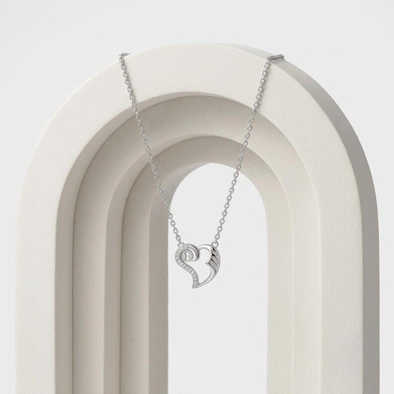 A silver heart-shaped pendant necklace with a small wing detail, hanging from a delicate silver chain on a white arched display stand.