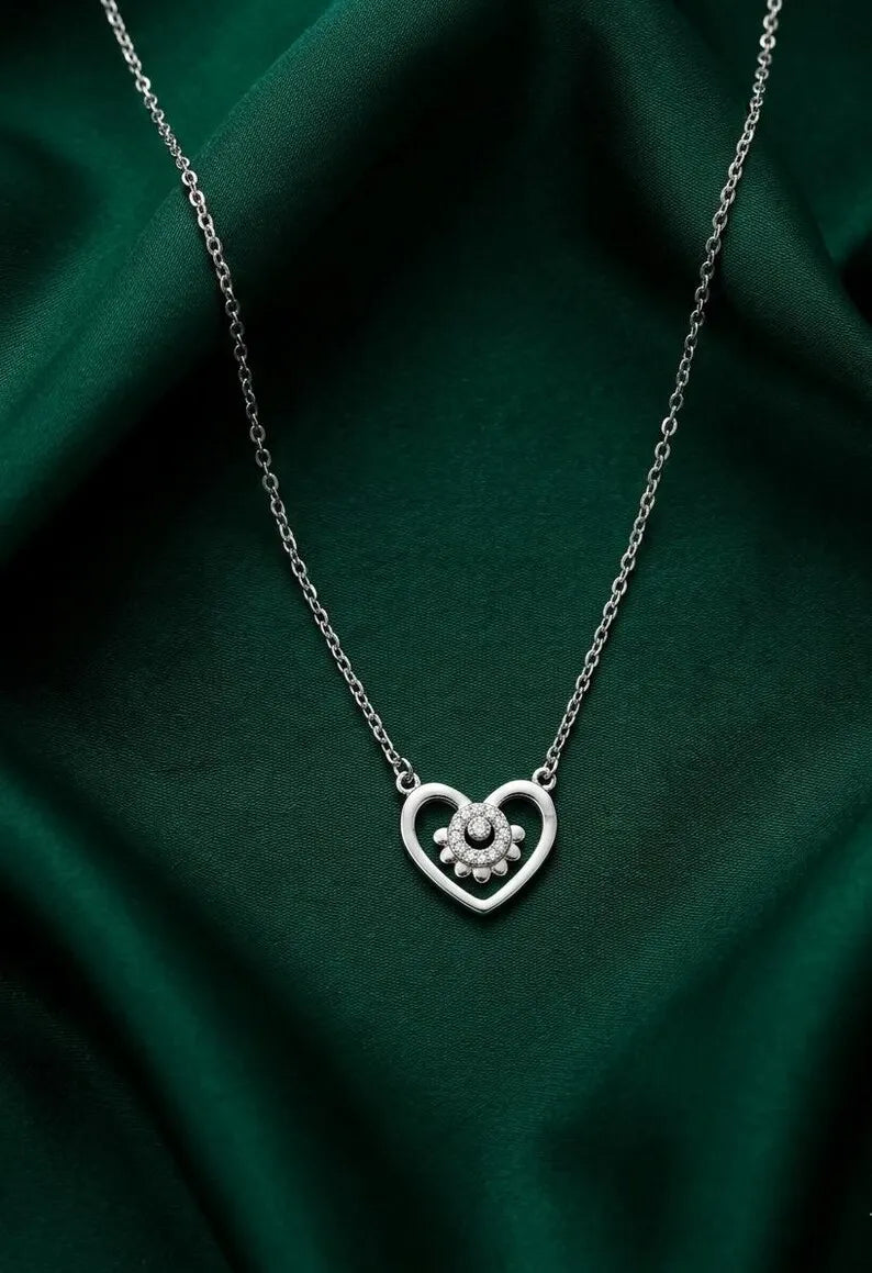 A silver heart-shaped pendant necklace with a small floral cluster in the center, displayed on draped dark green satin fabric.
