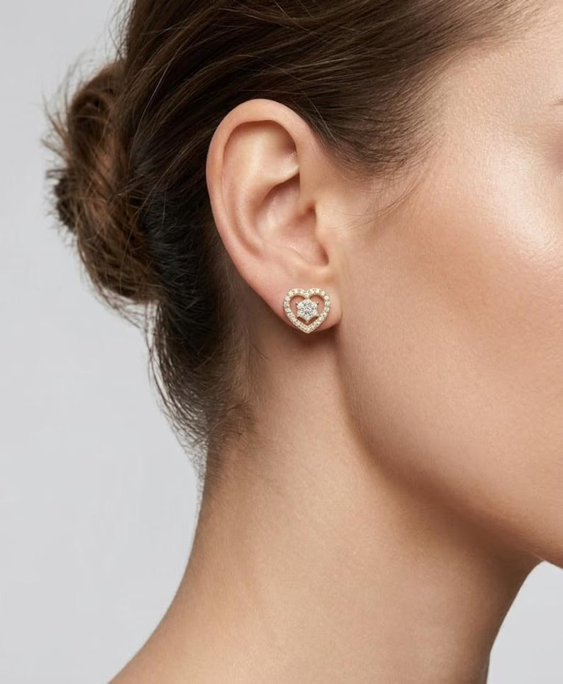 A close-up of a model's ear wearing a small heart-shaped stud earring with a gold-tone outline and a clear crystal center, set against a neutral light gray background.
