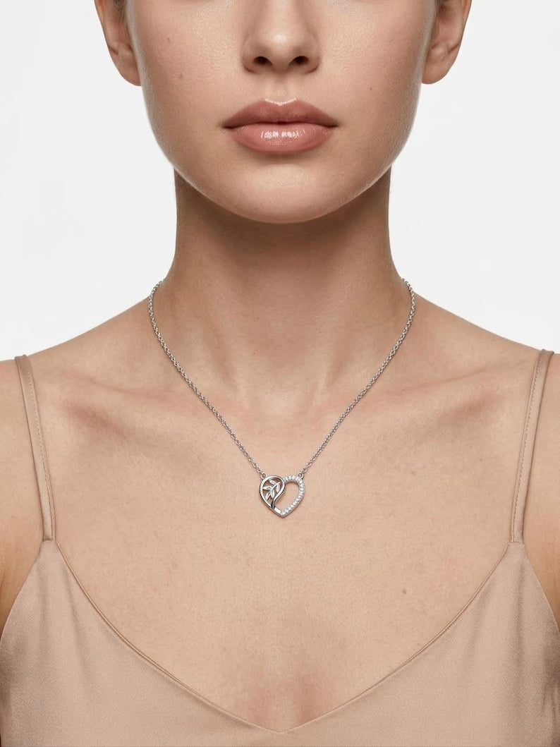 A close-up of a model wearing a delicate silver chain necklace with a heart-shaped pendant featuring a small silver cross in the center, styled with a beige camisole top against a light gray background.