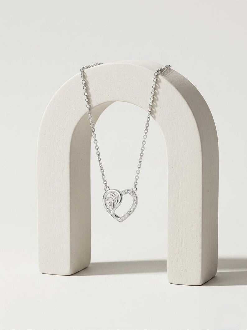 A silver heart-shaped pendant necklace with a pavé-set diamond center, draped over a minimalist white wooden arch display on a light beige background.