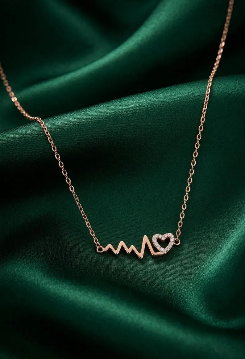 A rose gold necklace with a heartbeat line and heart pendant, displayed on draped emerald green satin fabric.
