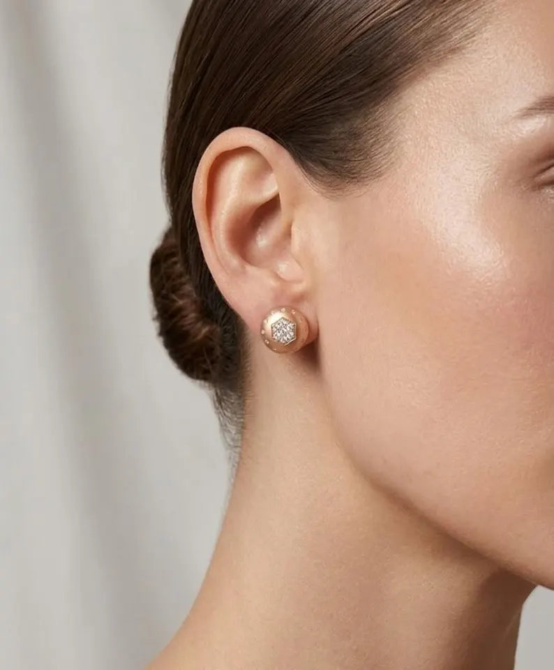 A close-up of a model's ear wearing a small round stud earring with a textured rose gold surface and clear stones.