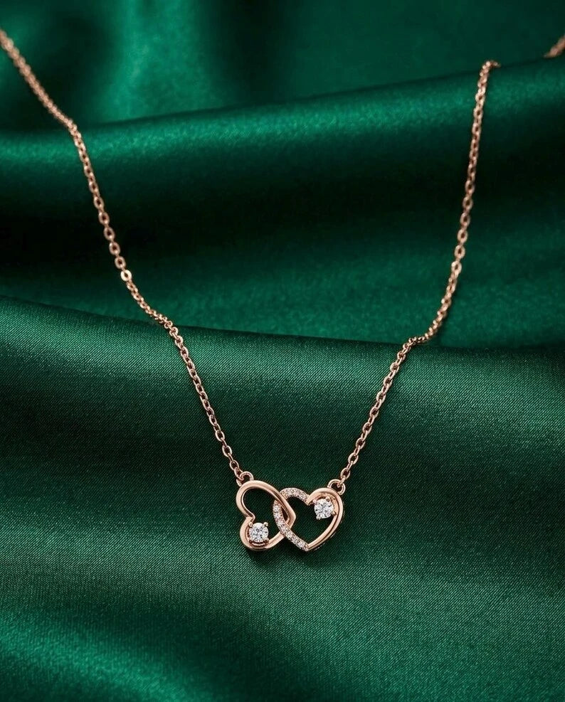 A rose gold necklace with two interlocking open heart pendants, one adorned with a small clear gemstone, displayed on a draped dark green satin fabric.
