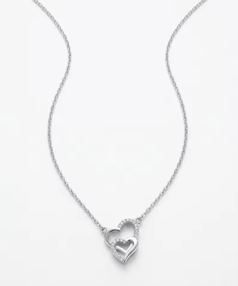 A silver chain necklace featuring a double-heart pendant, with one heart fully visible and the other partially enclosed by a halo of small clear stones, set against a plain white background.