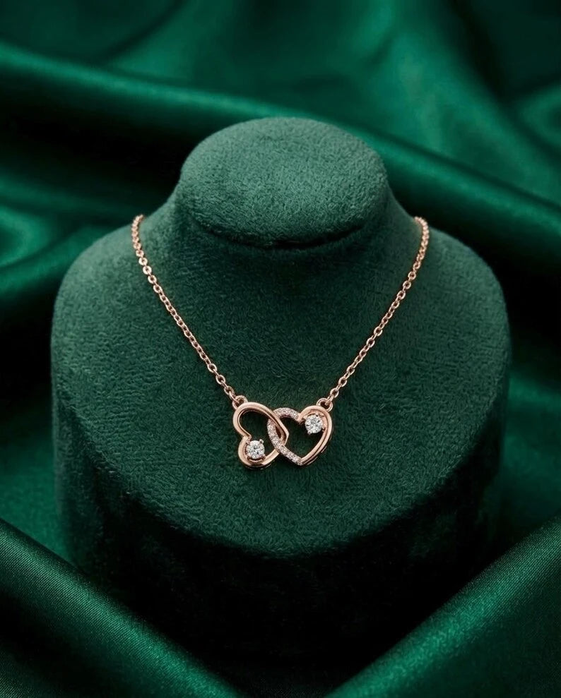 A rose gold necklace with two interlocking heart pendants, one set with clear stones, displayed on a dark green velvet jewelry bust against a matching green satin background.