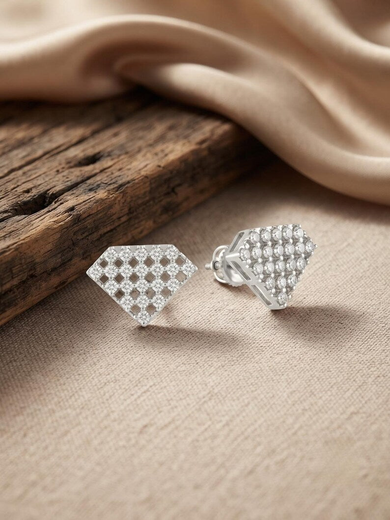 A pair of silver geometric stud earrings covered in small clear stones, displayed on a beige fabric surface next to a rustic wooden block and draped satin.