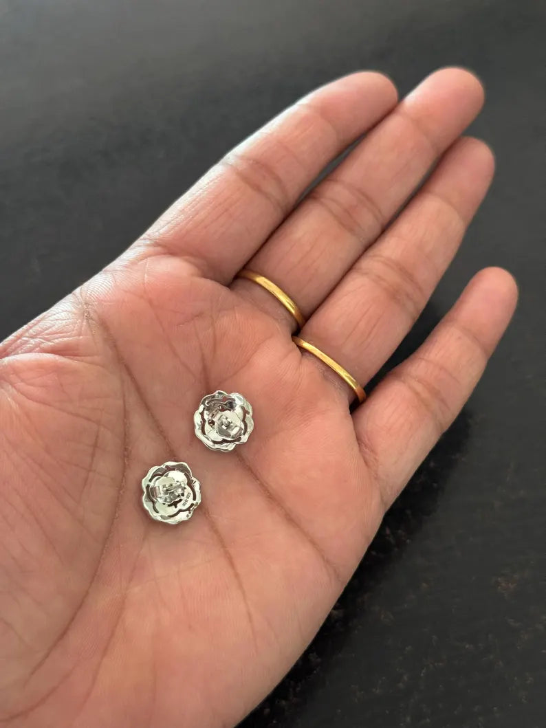 Two small silver-toned flower-shaped stud earrings resting on an open hand, with two gold rings worn on the fingers against a dark textured background.