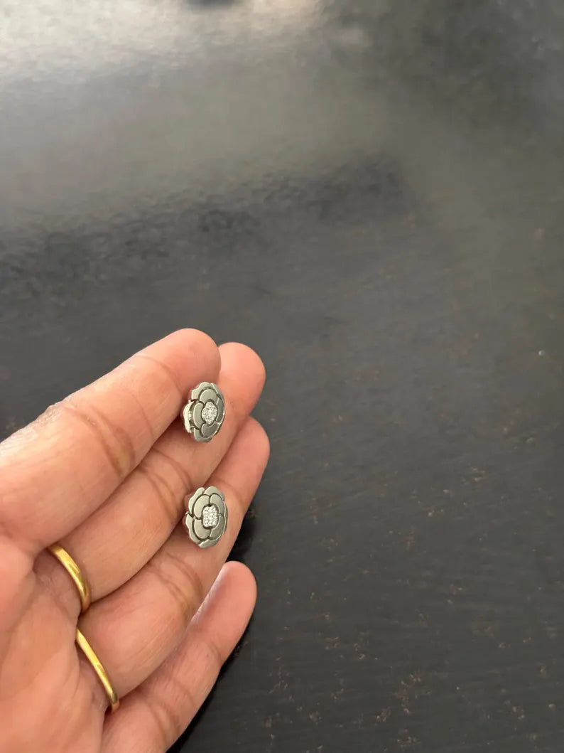 A close-up of a hand holding two small silver-toned flower-shaped earrings against a dark gray background.