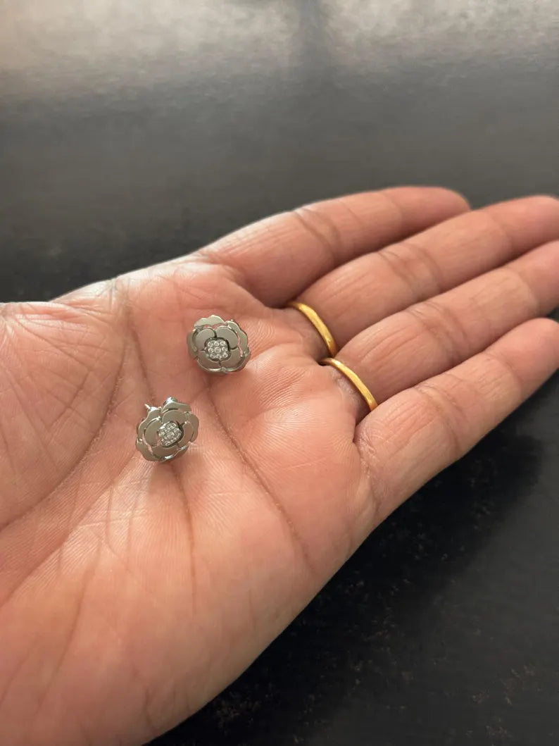 A close-up of a person's hand holding two small silver-toned flower-shaped earrings, with two gold rings on the fingers, against a dark gray background.