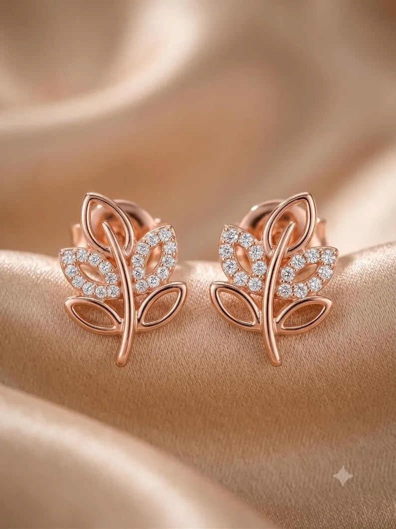 A pair of rose gold leaf-shaped stud earrings adorned with small clear pavé stones, displayed on a draped beige satin fabric background.