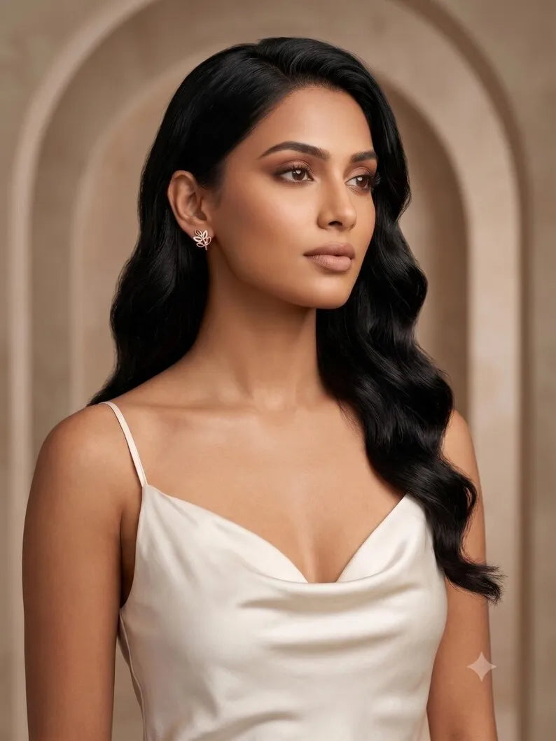 A woman with long, wavy black hair wearing a white spaghetti-strap top and a small silver stud earring, posed against a warm beige arched background.