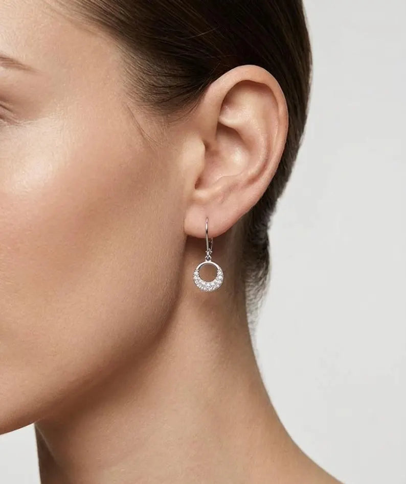 A close-up of a model's ear wearing a silver hoop earring adorned with small clear stones, set against a plain light gray background.