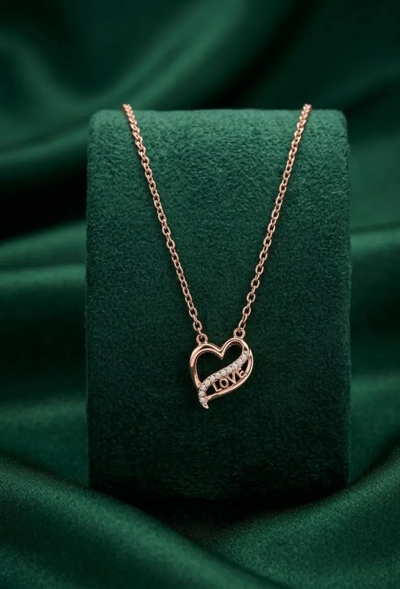 A rose gold heart-shaped pendant necklace with the word 'LOVE' in a small clear stone, displayed on a dark green velvet jewelry stand against a draped green satin background.