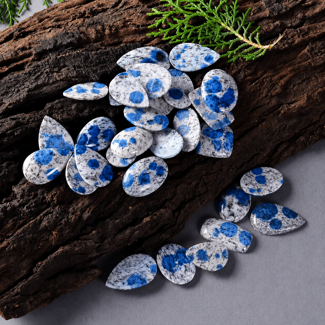Blue and white spotted teardrop-shaped gemstone pendants arranged on a piece of dark wood with a small green sprig, on a gray surface.