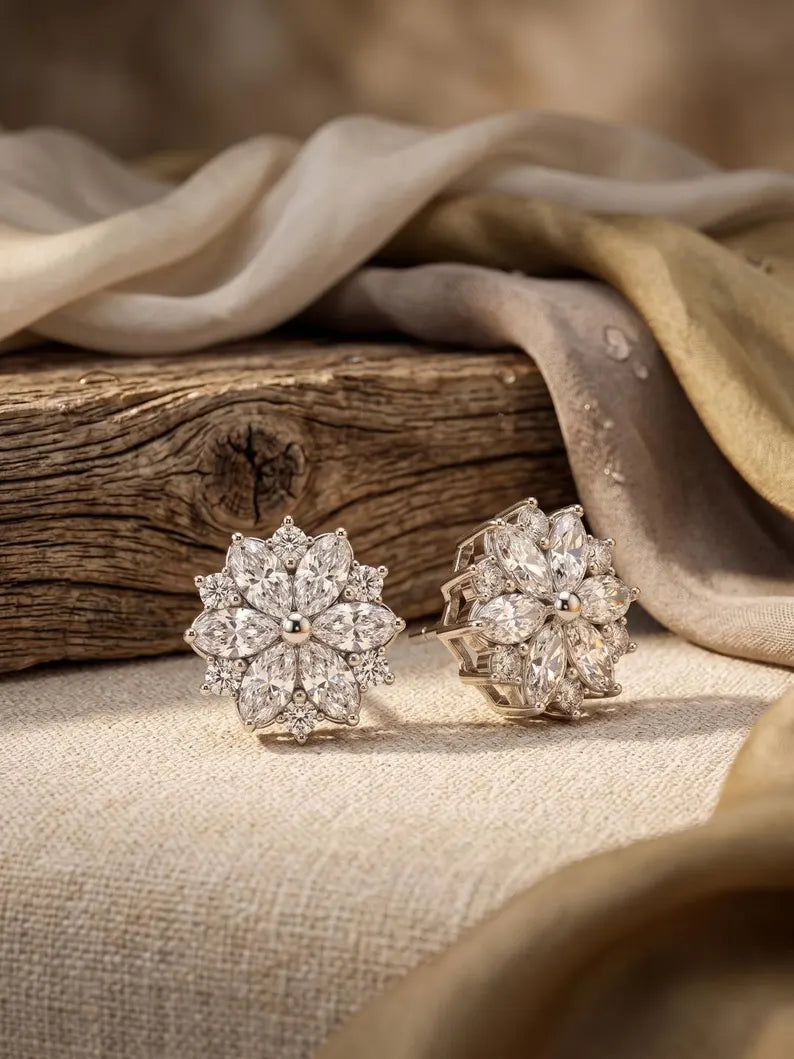 A pair of silver-toned stud earrings with intricate floral clusters of clear stones, displayed on a textured beige fabric next to a rustic wooden block with draped beige and gold fabric in the background.