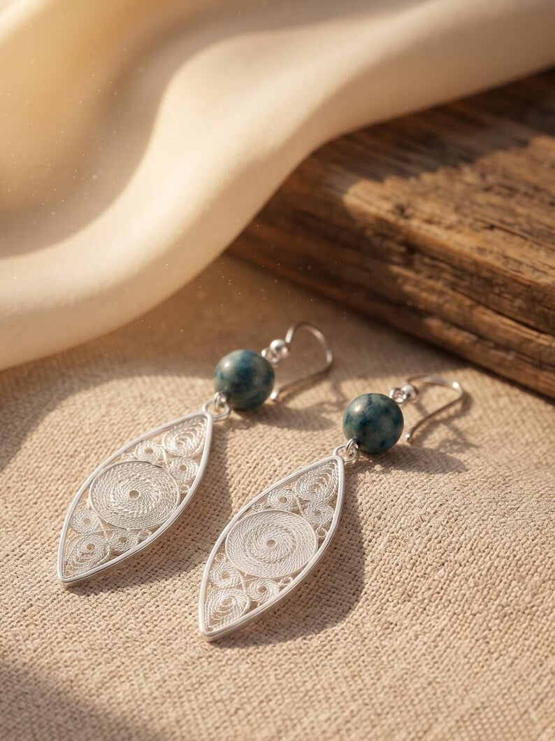 A pair of silver filigree dangle earrings with round dark green beads, displayed on a textured beige fabric next to a rustic wooden block.