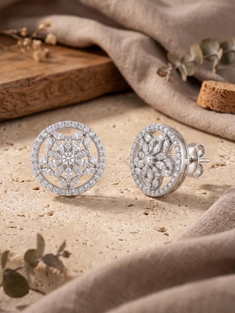 A pair of round silver stud earrings with intricate pavé crystal mandala designs, displayed on a textured beige stone surface with draped fabric and dried botanicals in the background.