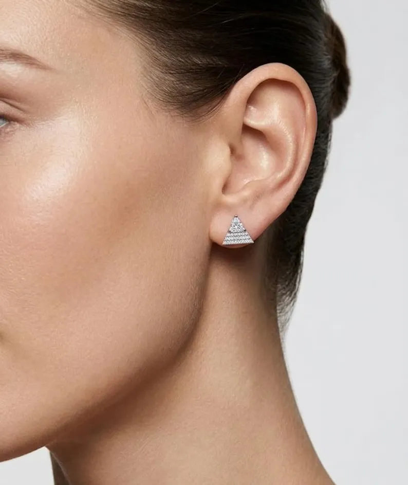 A close-up side profile of a woman wearing a small triangular stud earring with a silver-toned pyramid design set with clear stones, against a light gray background.