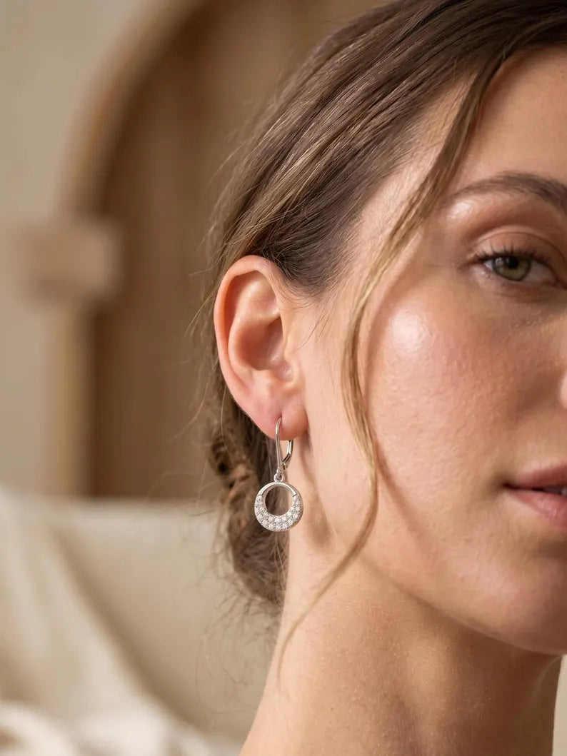 A close-up of a woman wearing a delicate silver hoop earring with a clear crystal border, set against a softly blurred warm beige background.
