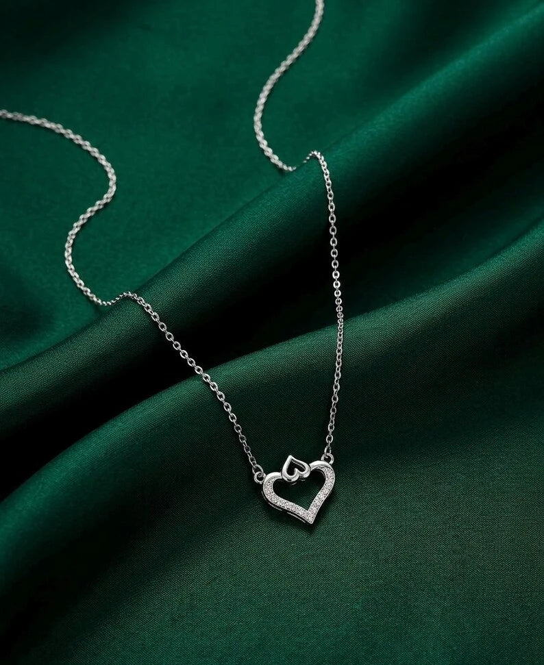 A silver heart-shaped pendant necklace with a small open heart design, displayed on a draped dark green satin fabric background.