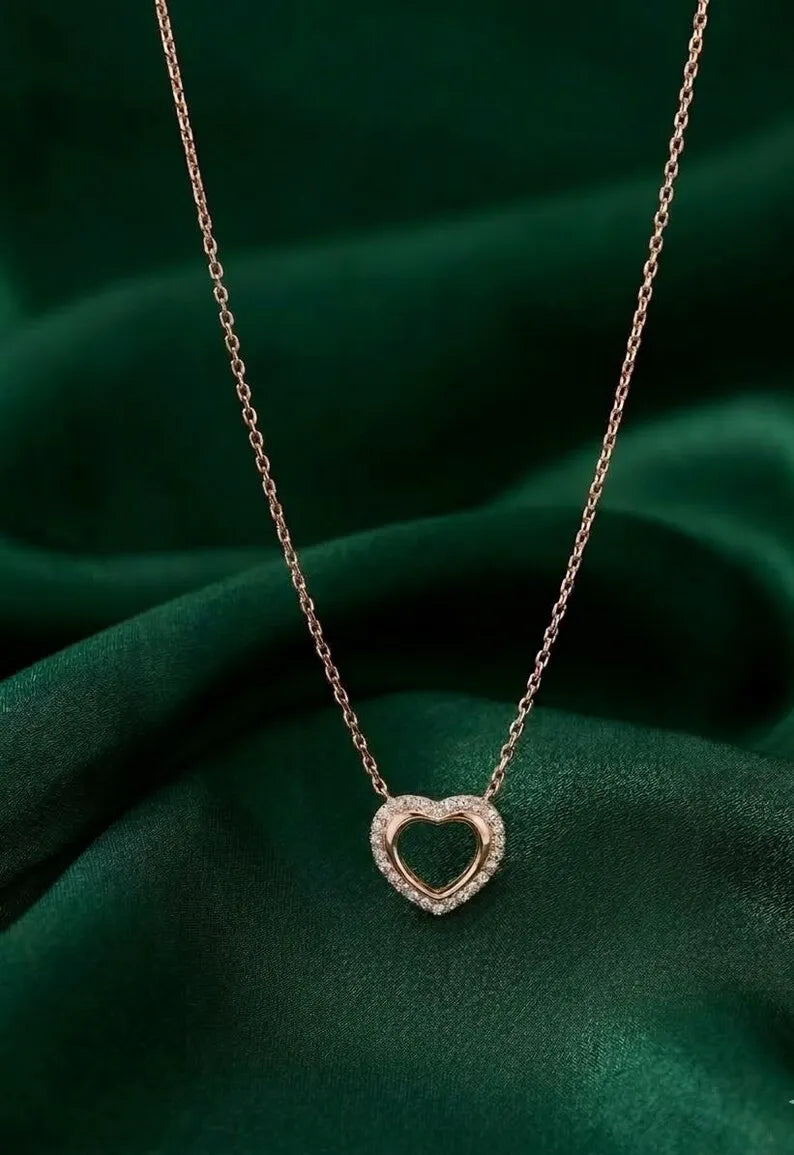 A delicate rose gold chain necklace with a small, double-heart pendant encrusted with clear stones, displayed on a draped dark green satin fabric.