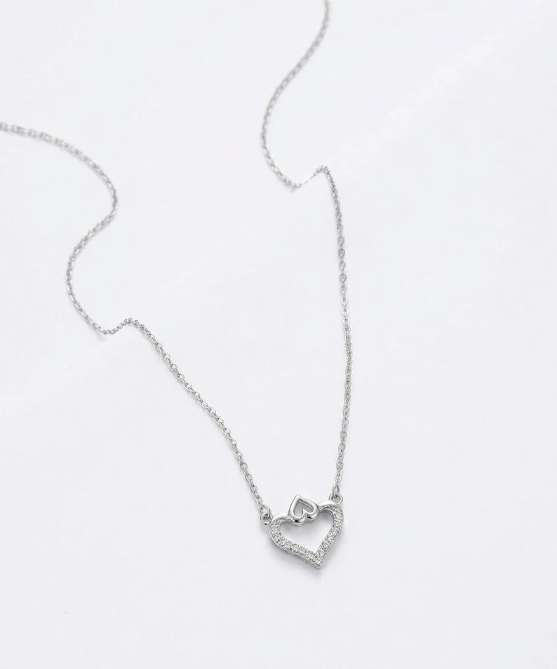 A delicate silver chain necklace featuring a small open heart pendant with a tiny heart inside, displayed on a clean white background.