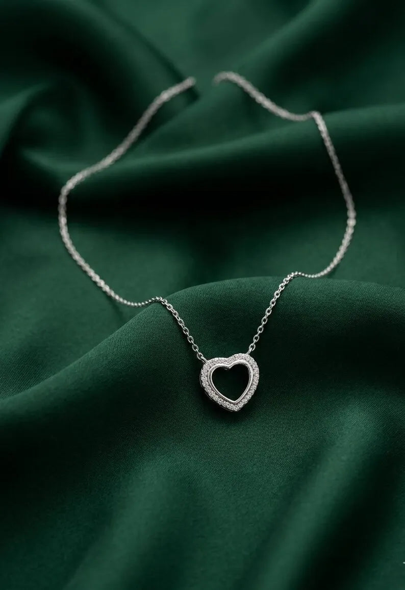 A silver heart-shaped pendant necklace with a delicate chain, displayed on draped dark green satin fabric.