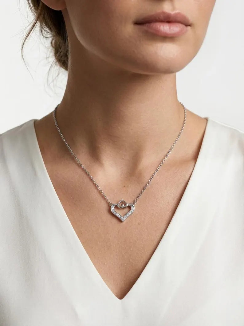 A close-up of a woman wearing a delicate silver heart-shaped pendant necklace with a small central ring detail, styled with a white V-neck top against a clean white background.