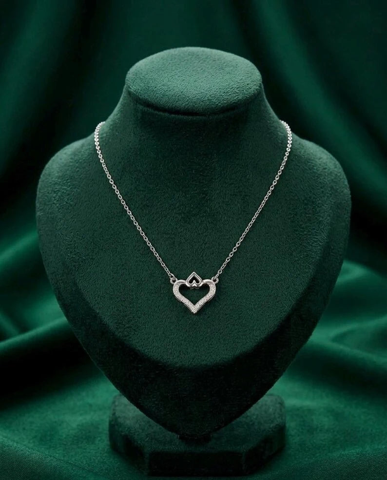A silver heart-shaped pendant necklace displayed on a dark green velvet jewelry bust against a matching green fabric backdrop.