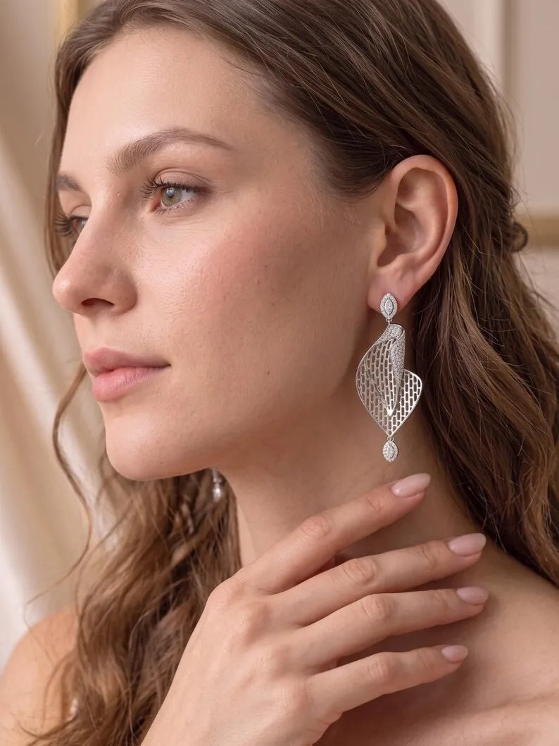 A close-up of a woman wearing a dangling silver earring with a textured, openwork leaf or petal design and a small teardrop charm.